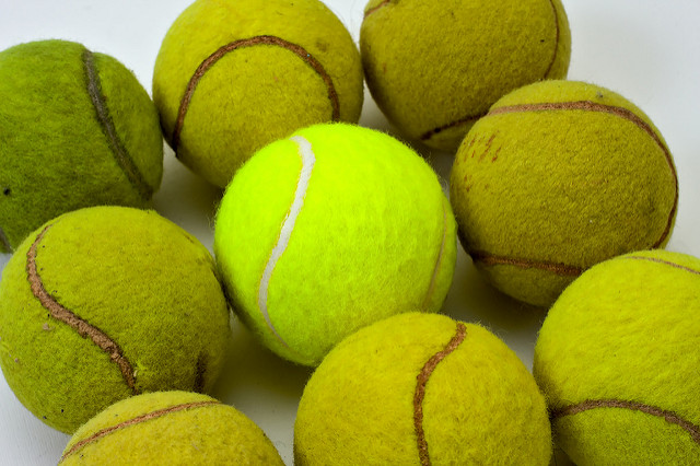 Brand new tennis ball with bright fluorescent green felt and white rubber band, surrounded by eight other used balls with duller, more washed out colors, deteriorated nap and dirt marks.
